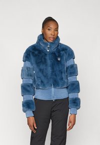 Casaco cropped de peles sintéticas em azul-escuro com mangas em malha. Gola alta e fecho frontal com zíper. Padrão texturizado com seções de tecido contrastantes.