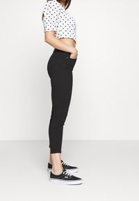 Svarta smala jeans med hög midja, framfickor och en smidig design, ihop med en vit topp med prickar och svarta sneakers.