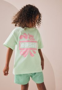 Tricou verde deschis cu un logo roz în formă de splash și textul „oasis”, asortat cu pantaloni scurți verzi în dungi. Hainele au croială lejeră și textură moale.