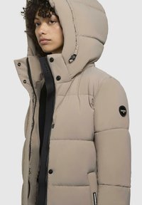 Beige pufferjakke med hætte, med en quiltet design, sorte accenter, trykknapper og et logopatch på venstre ærme.