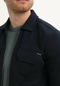 Chaqueta con cremallera en color navy, con cuello y dos bolsillos frontales, hecha de una tela suave. Debajo, se puede ver un suéter ligero de cuello redondo en color verde claro.