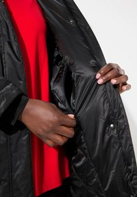 Giubbotto imbottito nero con texture lucida e chiusure a pressione, aperto per rivelare una camicia rossa sottostante. Mano che accede a una tasca interna.