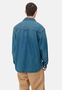 Camisa de denim en azul oscuro con un corte relajado. El diseño incluye un cuello, puños abotonados y un dobladillo curvado. Tejido texturizado con costuras visibles.