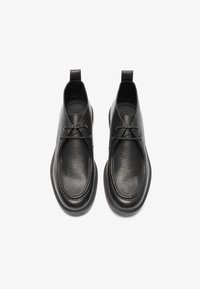 Zapatos de cuero negro con superficie texturizada, puntera redondeada, cordones minimalistas y tirador en el talón. Suela plana y lisa.