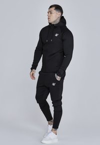 Czarny zestaw z bluzą na zamek i spodniami o dopasowanym kroju, wykonany z gładkiej tkaniny. Posiada logo na klatce piersiowej i z boku spodni. Białe sneakersy.