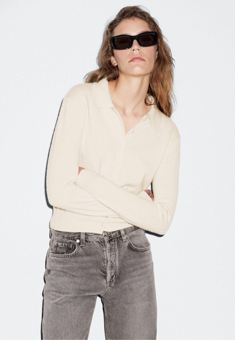 Cream-colored, long-sleeve polo top with a button-down front, paired with gray denim jeans. The fabric appears soft and slightly textured.