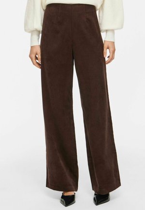 Femme portant un pantalon marron taille haute à jambes larges, un haut blanc à manches longues et des chaussures noires à bouts pointus, debout sur fond blanc.