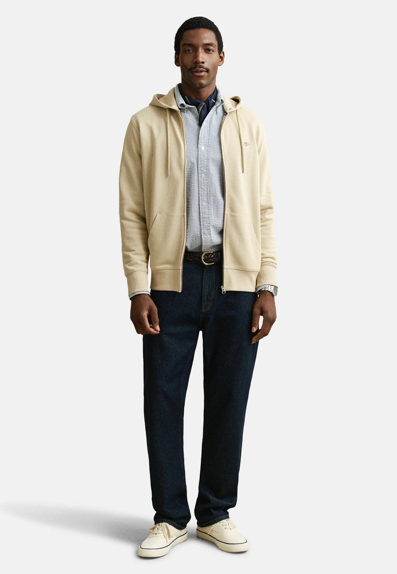 Mann trägt einen beigen Kapuzenpullover mit Reißverschluss über einem blau karierten Hemd, dunkle Jeans und weiße Sneaker, steht vor einem schlichten Hintergrund.