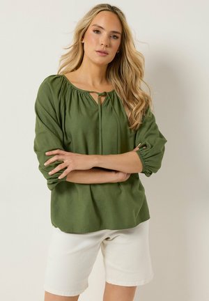Blonde vrouw met lang haar die een olijfgroene blouse met lange mouwen en witte korte broek draagt, staand met gekruiste armen tegen een effen achtergrond.