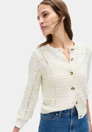 Frau trägt eine cremefarbene, strukturierte Strickjacke mit Schildpattknöpfen und blaue Jeans, schaut seitlich und hat die Haare über das Gesicht geweht.