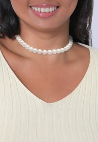 Collier ras du cou en perles blanches composé de perles rondes espacées de manière uniforme, porté autour du cou, associé à un haut à côtes légères.