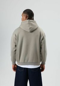 Sweat à capuche gris clair avec une coupe décontractée, poignets et ourlet côtelés. Porté sur une chemise blanche, associé à un jean large bleu foncé.