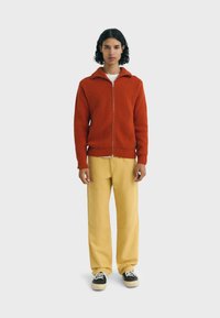 Oranje geribbelde jas met rits en hoge kraag, gedragen over een wit shirt. Gecombineerd met gele wijde broekspijpen en zwarte sneakers.