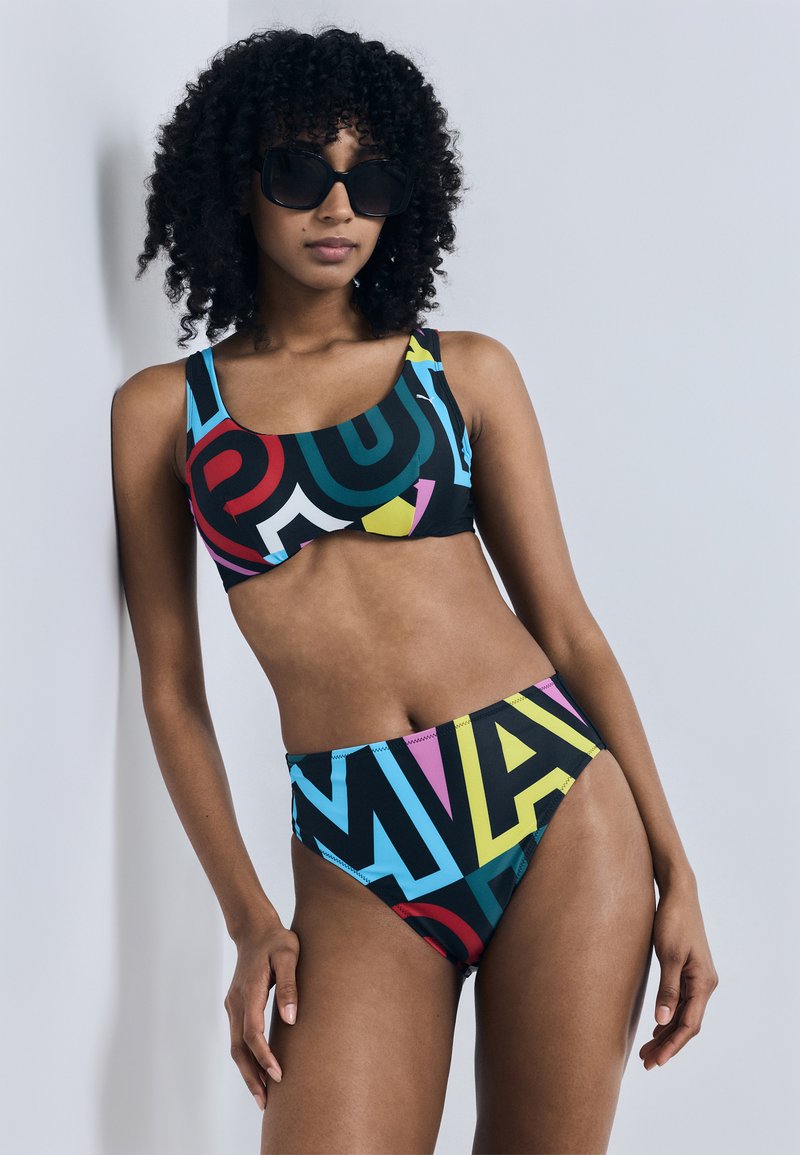 Γυναίκα με σγουρά μαλλιά και γυαλιά ηλίου, φοράει τοπ μπικίνι με πολύχρωμο γεωμετρικό μοτίβο και ψηλόμεσο κάτω μέρος, σε απλό φόντο.