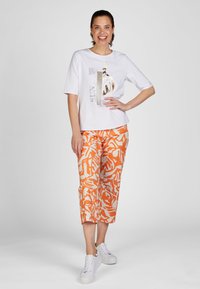 Weißes T-Shirt mit kurzen Ärmeln, das metallisch goldene Schriftzüge zeigt, kombiniert mit orangefarbenen, knöchellangen Hosen, die ein auffälliges abstraktes weißes Muster aufweisen. Weiße Sneakers.