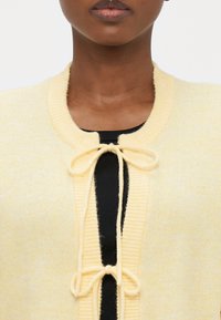 Yellow cardigan with a round neckline, featuring a tie closure, ribbed texture, and a soft, lightweight material over a black garment.