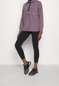 Pullover-Jacke mit mehrfarbigem Muster, hohem Kragen und halbem Reißverschluss, kombiniert mit schwarzen Leggings und schwarzen Sportschuhen.