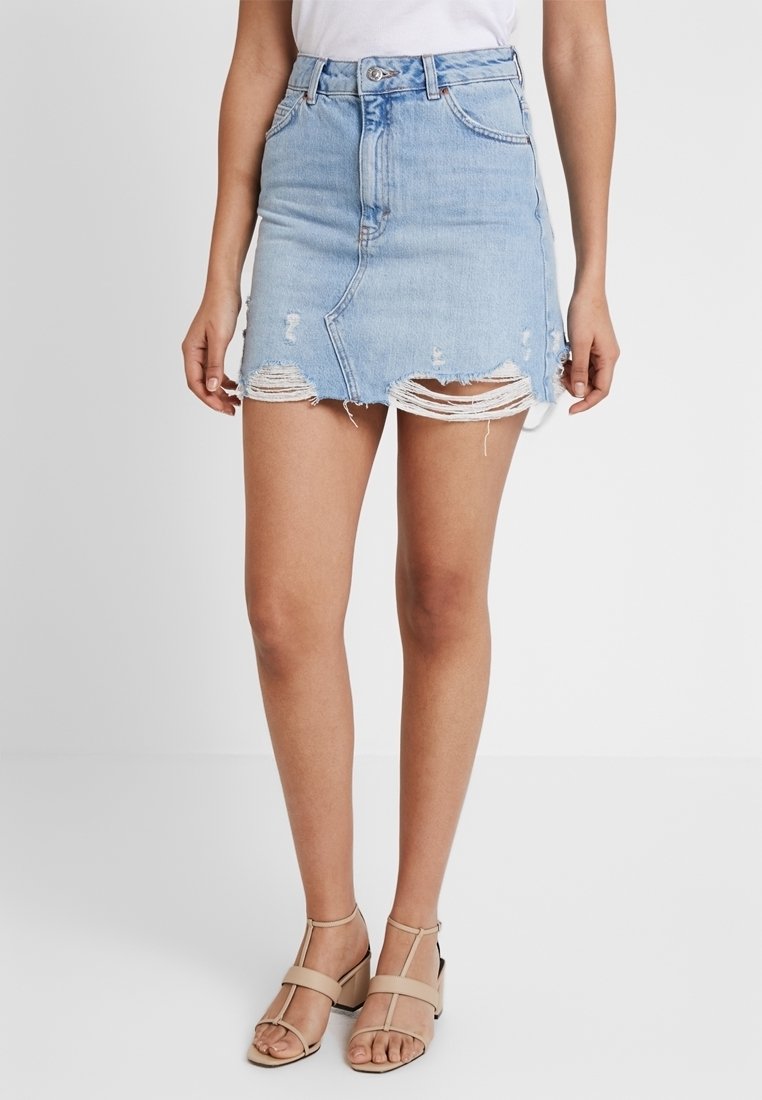 Femme portant une mini-jupe en denim bleu clair délavé avec un ourlet brut et des sandales beige à talon bloc ouvertes à l'avant, debout devant un fond uni.
