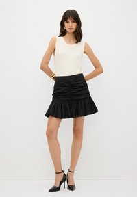 Bílý žebrovaný top na ramínka ve spojení s černou mini sukní s řasením a oblíbeným lemem. Modelka má na sobě černé lakované lodičky a zlaté doplňky.