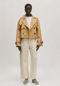 Afgeknotte tan trenchcoat met grote knopen, gecombineerd met hoge crèmekleurige wijde jeans en zwart-wit gestreepte instapschoenen.