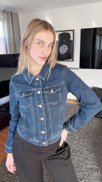 Νεαρό άτομο με ίσια ξανθά μαλλιά που φοράει μπλε τζιν μπουφάν και μαύρο παντελόνι, στέκεται μέσα σε εσωτερικό χώρο με το χέρι στη μέση.