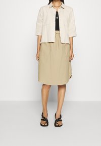 Beige cropped takki, musta yläosa, beige kiristyskuminauhalla varustettu hame ja mustat sandaalit. Huomattavina yksityiskohtina ovat napit edessä ja kaksi sivutaskua.