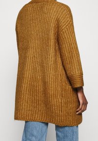 Lång, oversized cardigan i en djup senapsgul färg, med en ribbad textur och trekvartslängd på ärmarna. Bakifrån syns en rundad nederkant.