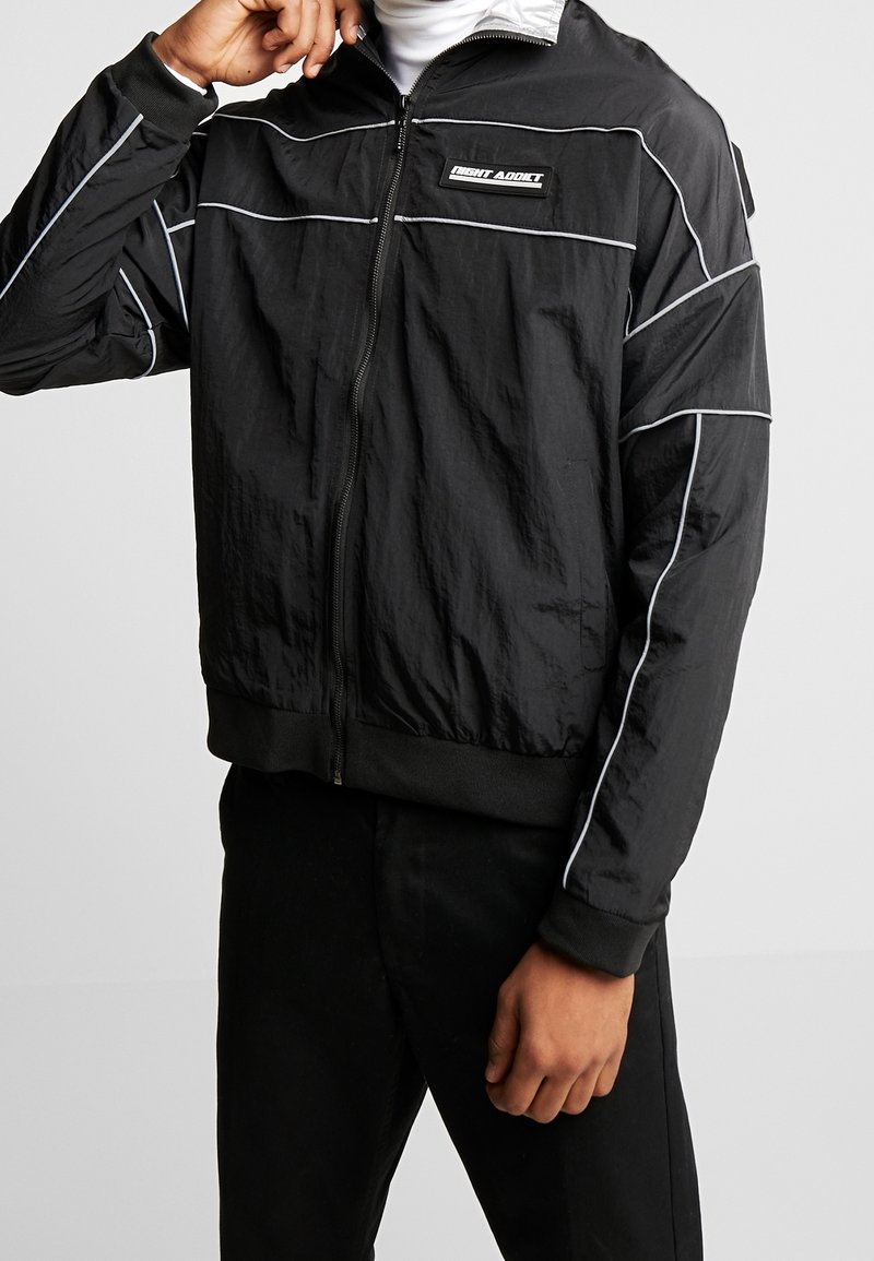 Veste noire à fermeture éclair avec passepoil blanc et écusson "Light Addict" sur la poitrine, portée sur un col roulé blanc, main près du col.