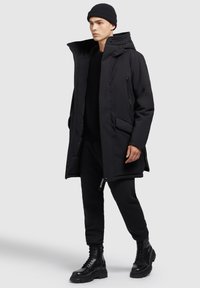 Sort parka med høj krave, lynlåslommer og et elegant design. Båret over en sort sweater og sweatpants; samlet med sorte snørebåndsstøvler.