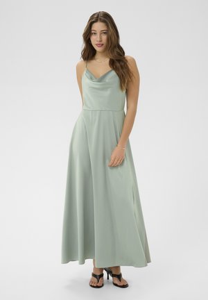 Donna con capelli lunghi che indossa un maxi abito senza maniche in raso verde pastello e sandali neri con tacco, in piedi davanti a uno sfondo uniforme.