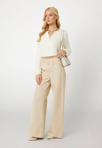 Witte donzige cropped cardigan, beige wijde broek en pumps met een spitse neus. Accessoires omvatten een kleine tas met kettingdetail.