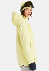 Gele geïsoleerde jas met een capuchon, voorzien van een groot voorvak, ritssluiting en zwarte manchetten over lichtgewicht handschoenen.
