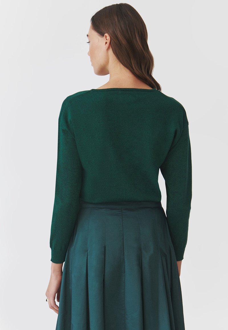 Grüner, metallic glänzender Strickpullover mit langen Ärmeln und weitem Auschnitt, kombiniert mit einem plissierten dunkelgrünen Rock, der eine glatte Textur und einen Kontrast zeigt.