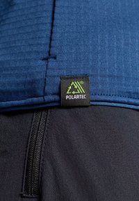 Tecido Polartec azul-marinho com textura canelada, apresentando uma etiqueta preta com logo verde. Vista de perto de um bolso com fecho éclair em material mais escuro.