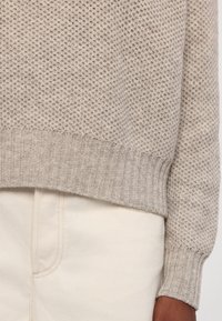 Poignet et ourlet en tricot beige texturé d'un pull, sur un pantalon couleur crème, avec une partie visible de la main d'une personne à la peau brune à droite.