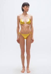 Γυναίκα που στέκεται ξυπόλητη φορά ένα λαμπερό χρυσό μπικίνι δύο τεμαχίων, κοιτάζοντας ευθεία μπροστά με το κεφάλι γυρισμένο ελαφρώς στο πλάι, σε ένα απλό λευκό φόντο.