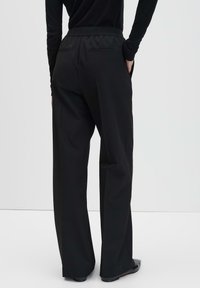 Personne portant un pantalon noir taille haute à jambes larges et un haut noir à manches longues, se tenant les mains dans les poches contre un fond clair.