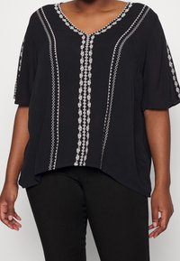 Svart topp med V-ringning, korta ärmar och vita broderade vertikala mönster längs framsidan. Tillverkad av lättviktigt tyg, avslappnad passform.