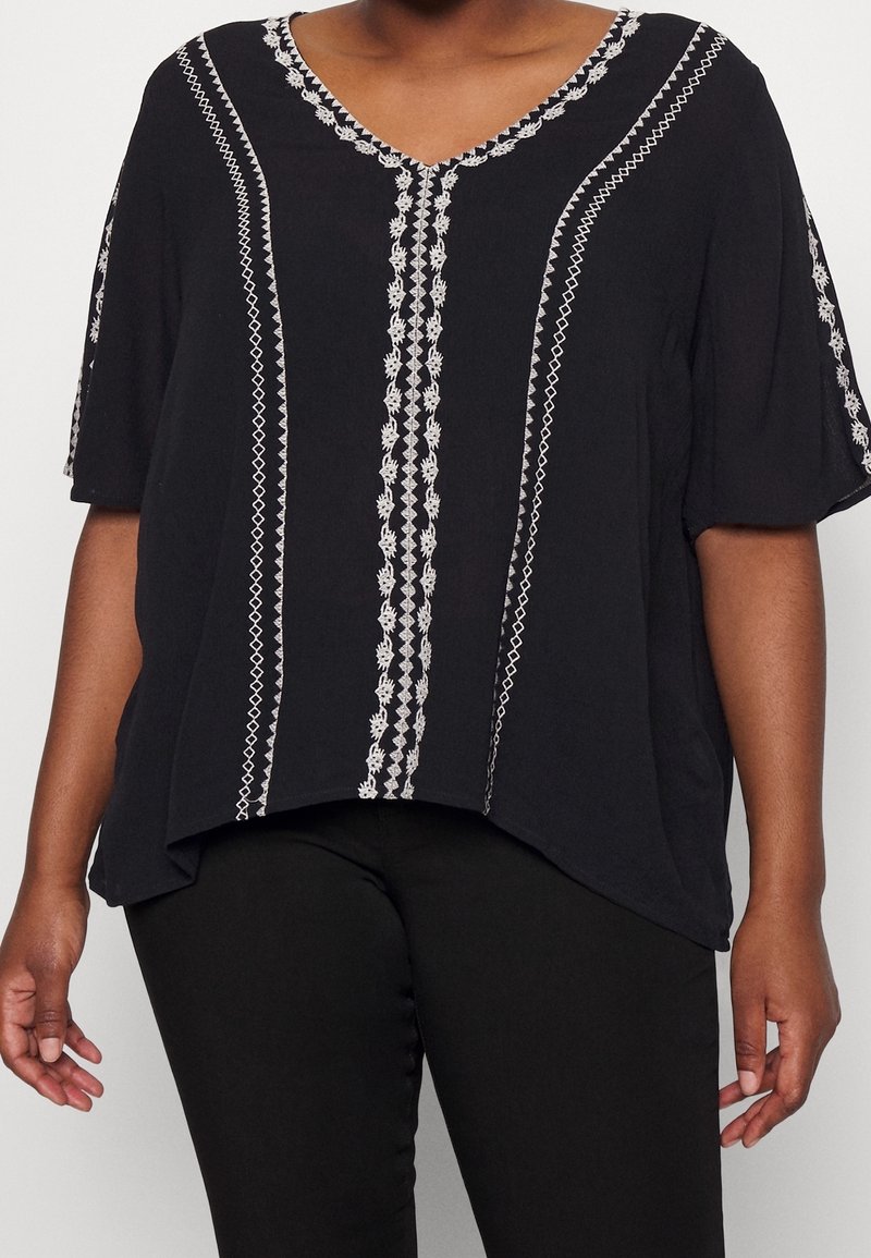Svart topp med V-ringning, korta ärmar och vita broderade vertikala mönster längs framsidan. Tillverkad av lättviktigt tyg, avslappnad passform.