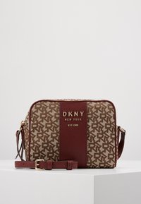 DKNY axelremsväska med ett mönster i tan och brunt logotyp, guldfärgad hårdvara och en vinröd läderdetalj. Har justerbar rem och rektangulär form.