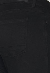 Detailní záběr na černé džíny, který ukazuje zadní kapsu a jemné šití na pravé straně.