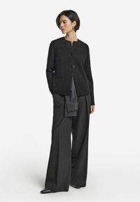 Sort strikket cardigan med rund hals, frontknapper og to lommer, båret over grå stribede bukser med vidde.