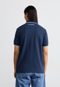 Marineblå polo med hvit kant på krage og ermer. Glatt stoff, logo detalj bak, og avslappet passform. Brukt med lyseblå jeans.