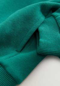 Tissu tricot teal avec des coutures visibles et un poignet côtelé replié sur un vêtement à manches longues, sur un fond clair.
