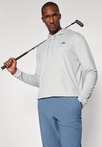 Sweatshirt capuz cinza claro com bolso frontal e logotipo preto, combinado com calças de golfe azuis estampadas. Homem segura um taco de golfe.