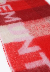 Rød og pink uldsjaal med et patchworkdesign i varierede nuancer og teksturer, herunder fuzzy områder og blokke med bogstaver.