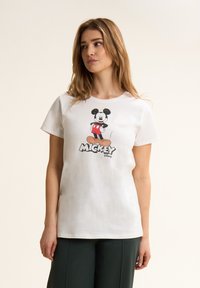 Camiseta de algodón blanca con un gráfico de Mickey Mouse en pantalones cortos rojos, con detalles en negro y la palabra "MICKEY" en letras gruesas.