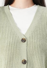 Zbliżenie osoby noszącej jasnozielony dziergany kardigan z dwoma brązowymi guzikami na białej prążkowanej koszuli.