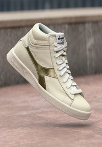 Baskets montantes en cuir crème avec un accent doré métallisé. Elles présentent des lacets blancs, une semelle texturée et un logo sur la languette.