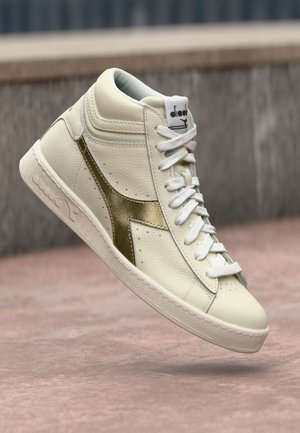 Baskets montantes en cuir crème avec un accent doré métallisé. Elles présentent des lacets blancs, une semelle texturée et un logo sur la languette.